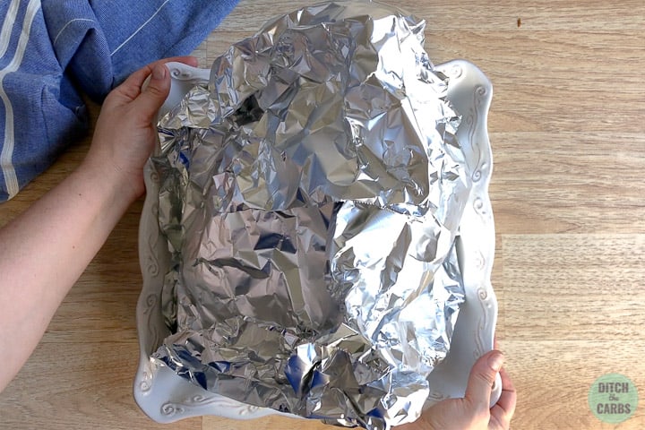 Keto ham wrapped in foil sitting in a white baking dish. Hands are lifting the dish to place in the oven.