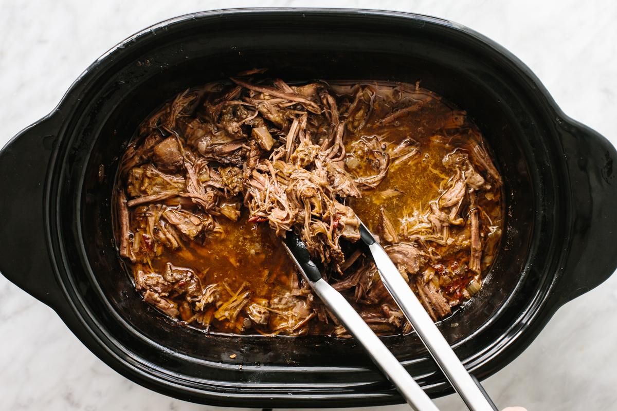 Picking up beef barbacoa in a slow cooker with tongs.