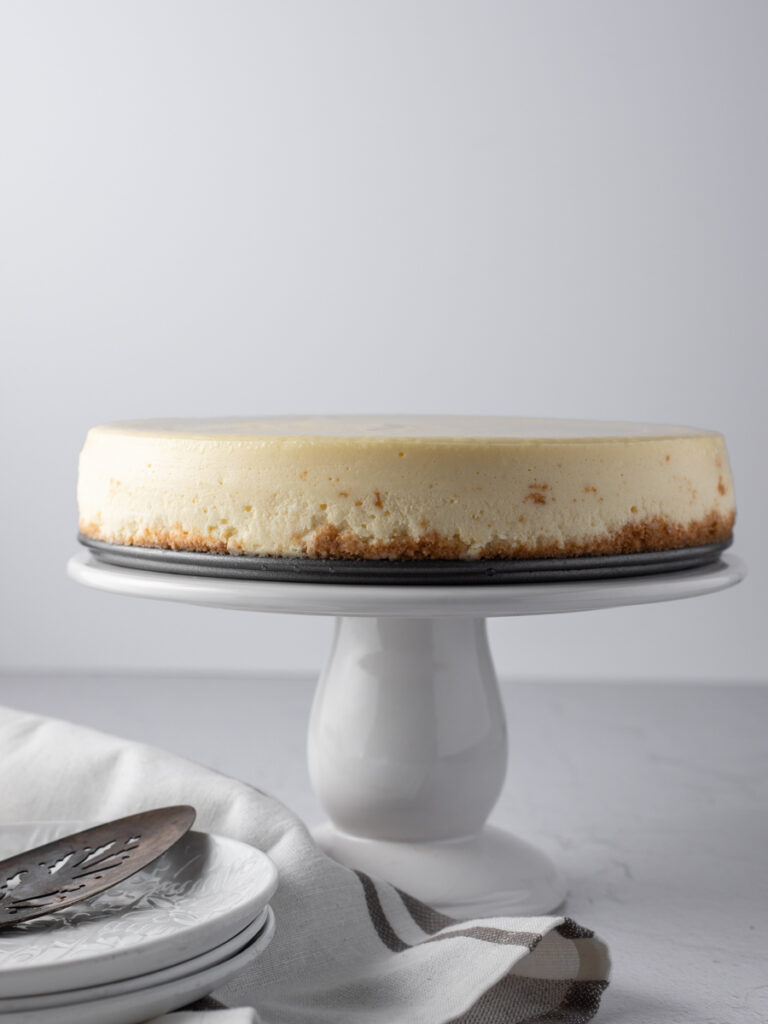 A whole New York cheesecake on a white cake stand.