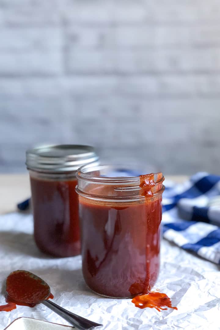 Two jars of keto BBQ sauce. One jar is opened with a drip of BBQ sauce sliding down the jar. A spoonful of BBQ sauce is resting on a the table to the left.