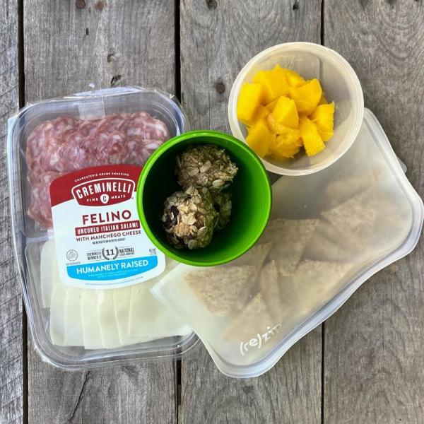 Packed school lunch that includes a charcuterie plate with salami and cheese, thin Triscuits, homemade Oatmeal Cookie Energy Bites, and mango. 