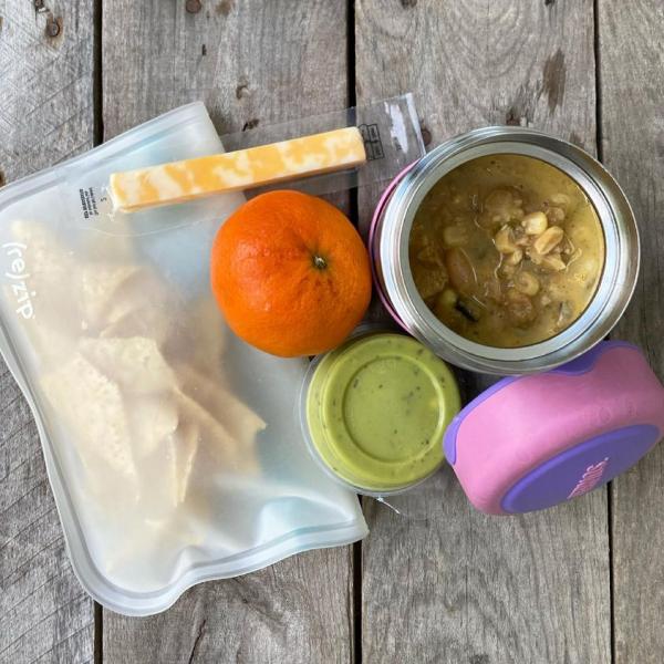 Packed school lunch that includes White Chicken Chili in a thermos, tortilla chips, guacamole, orange, and a cheese stick. 