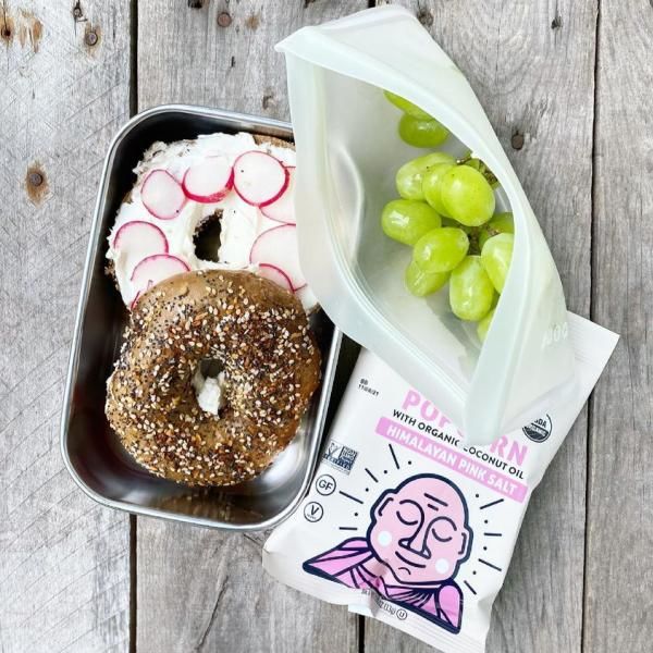Packed school lunch that includes a whole-wheat bagel topped with cream cheese and radishes, grapes, and popcorn. 