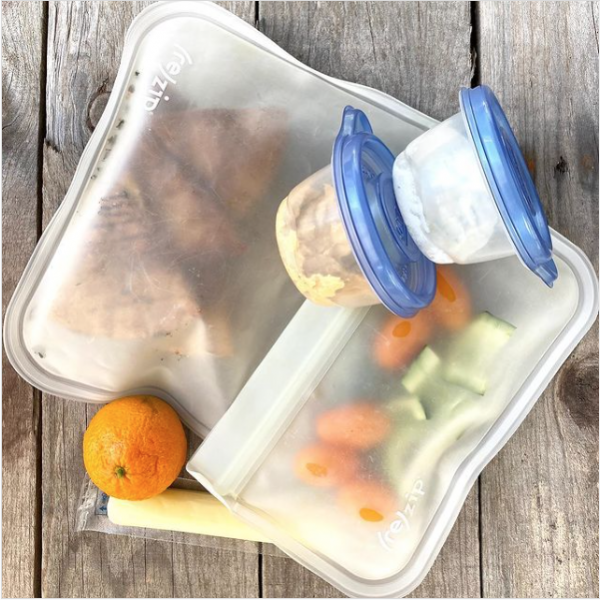Packed school lunch that includes baked pita chips, tomatoes, cucumbers, an orange, cheese stick, and tzatziki and hummus.  