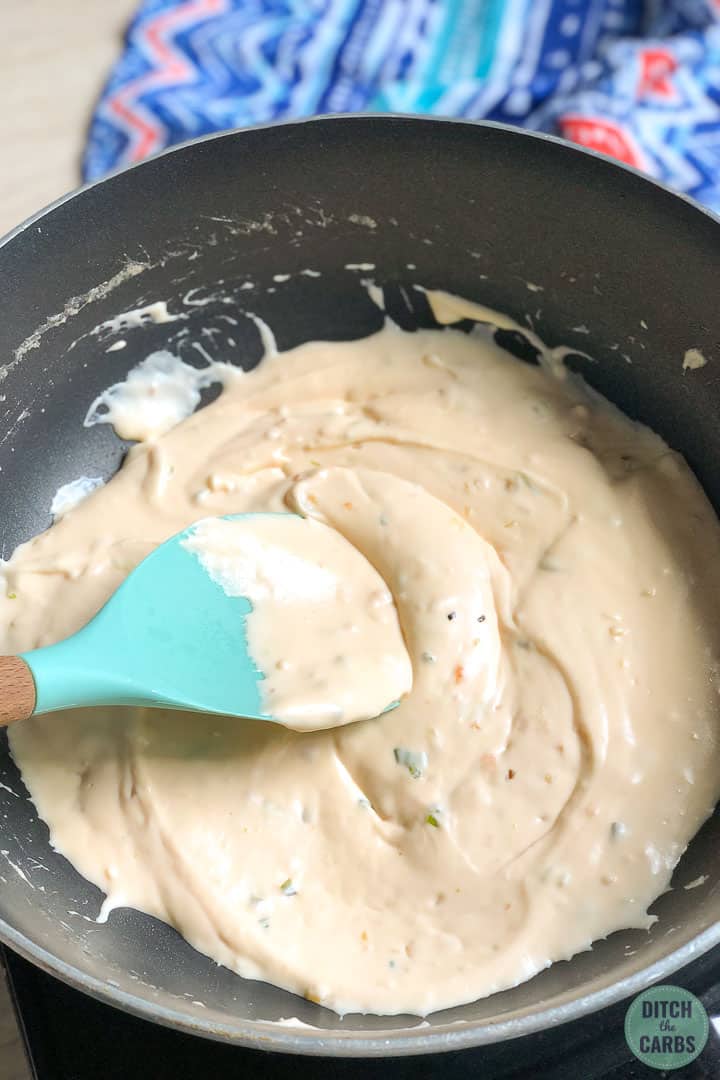 Creamy cheese sauce in a skillet being stirred by a light blue green rubber spatula for keto pulled pork mac and cheese.