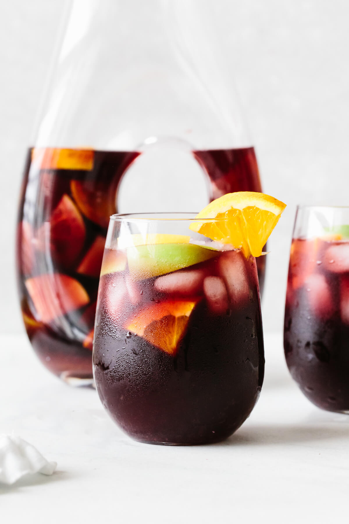 Red sangria in a glass with orange slice garnish and pitcher in the background.