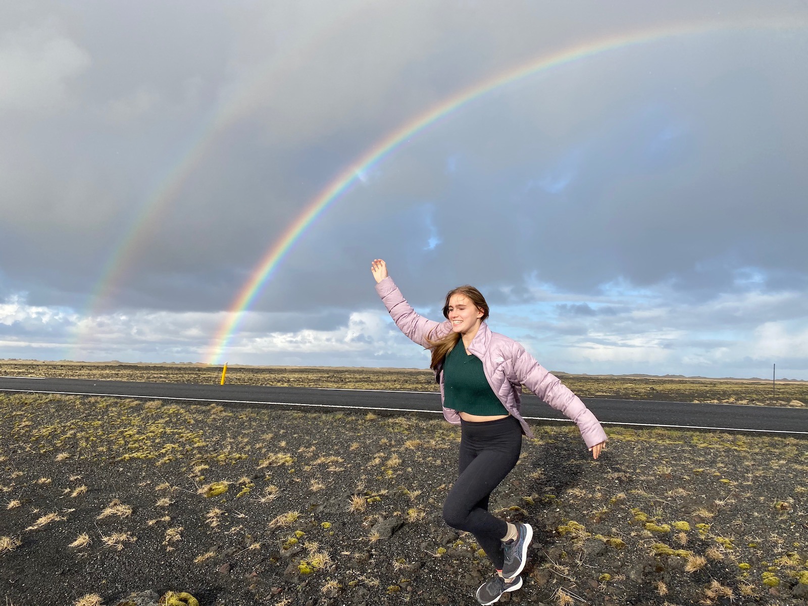 Teen girl in field with double rainbow in the background in Iceland. 