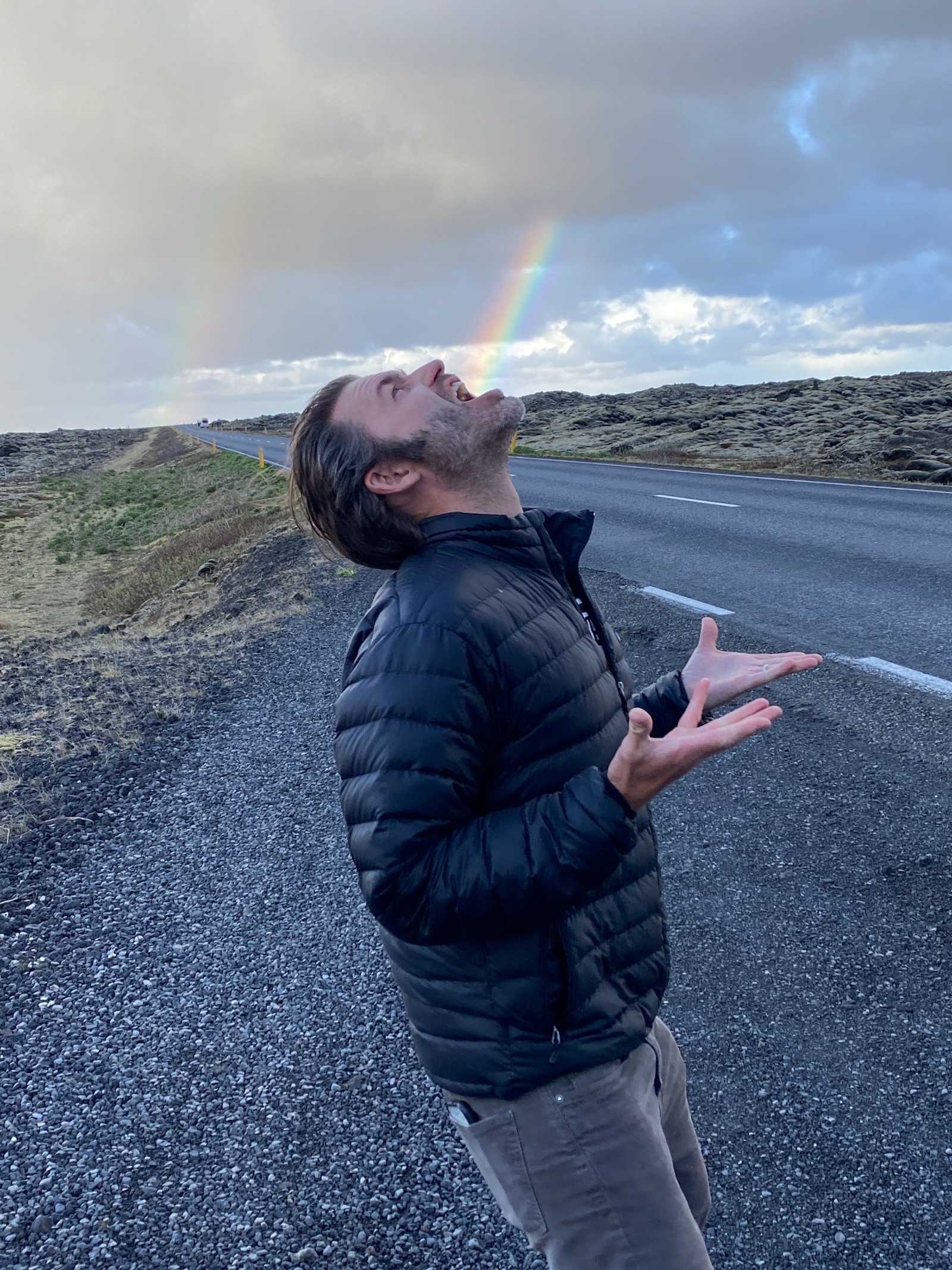 Father posing on the side of the road in Iceland with rainbows in the background. 