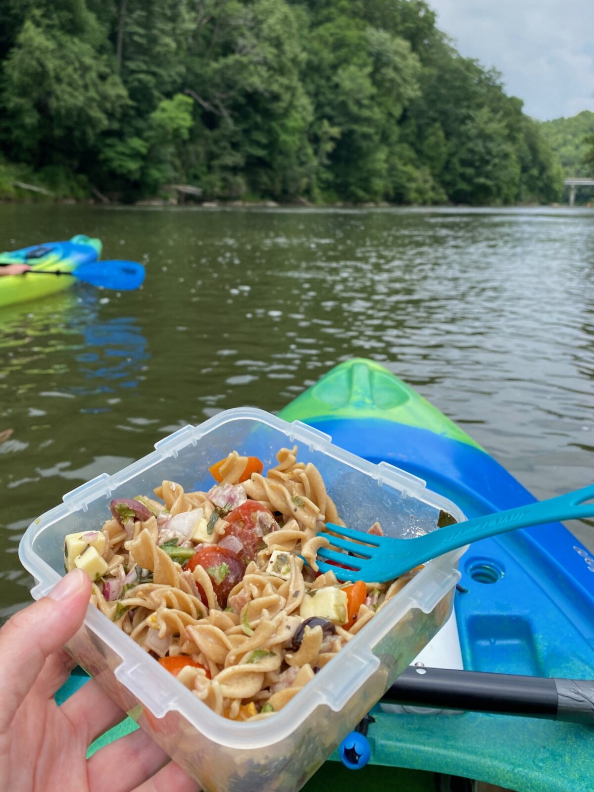 Holding a container of pasta salad while sitting in a kayak on the water from a persons viewpoint. 