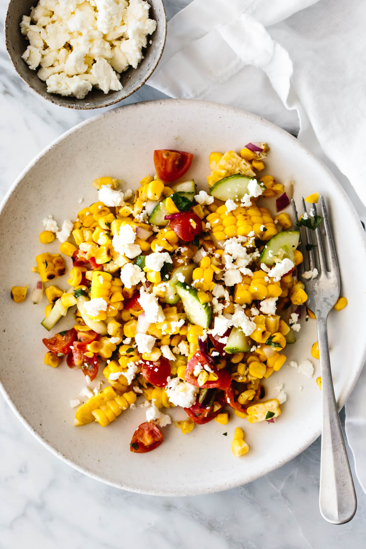 A white plate with grilled corn salad next to a bowl of feta cheese.