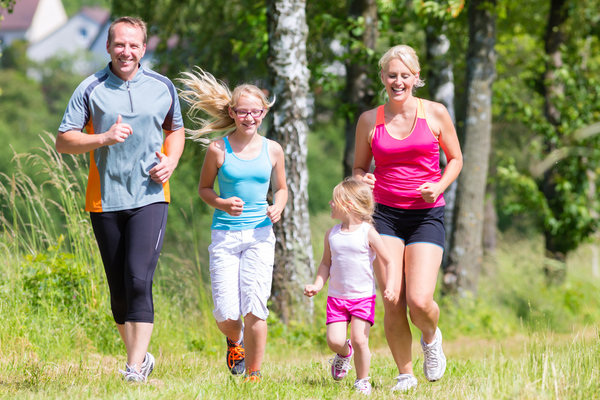Family of four including blonde mum wearing pink vest and black shorts, dad wearing blue tshirt and long length dark shorts, two blonde haired girls one wearing a light blue vest and white shorts and the other wearing a white vest and pink shorts. they are running together outside in a forest/field area and showing that it is good to get the whole family involved in fitness.
