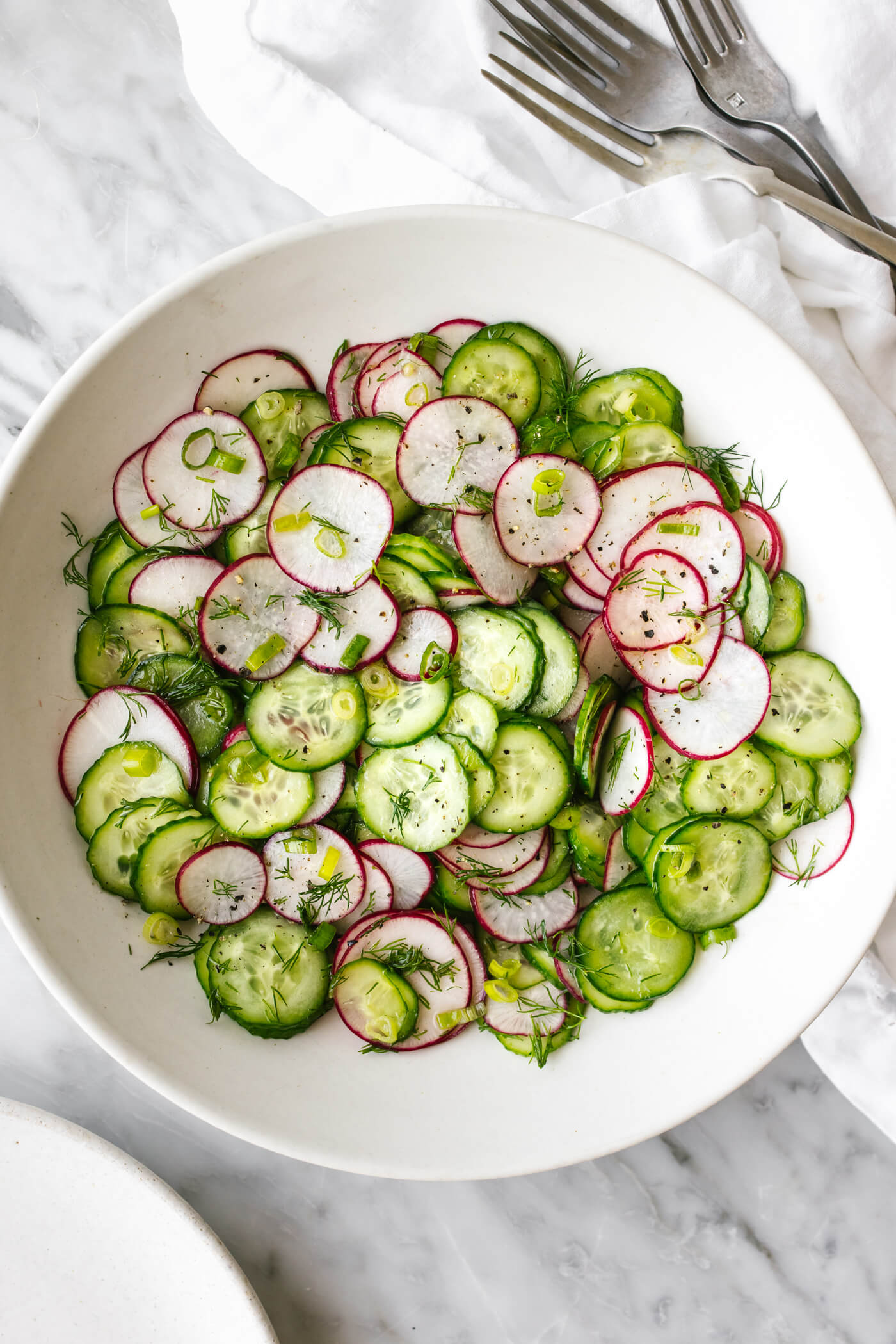 A white bowl of cucumber radish salad next to forks.