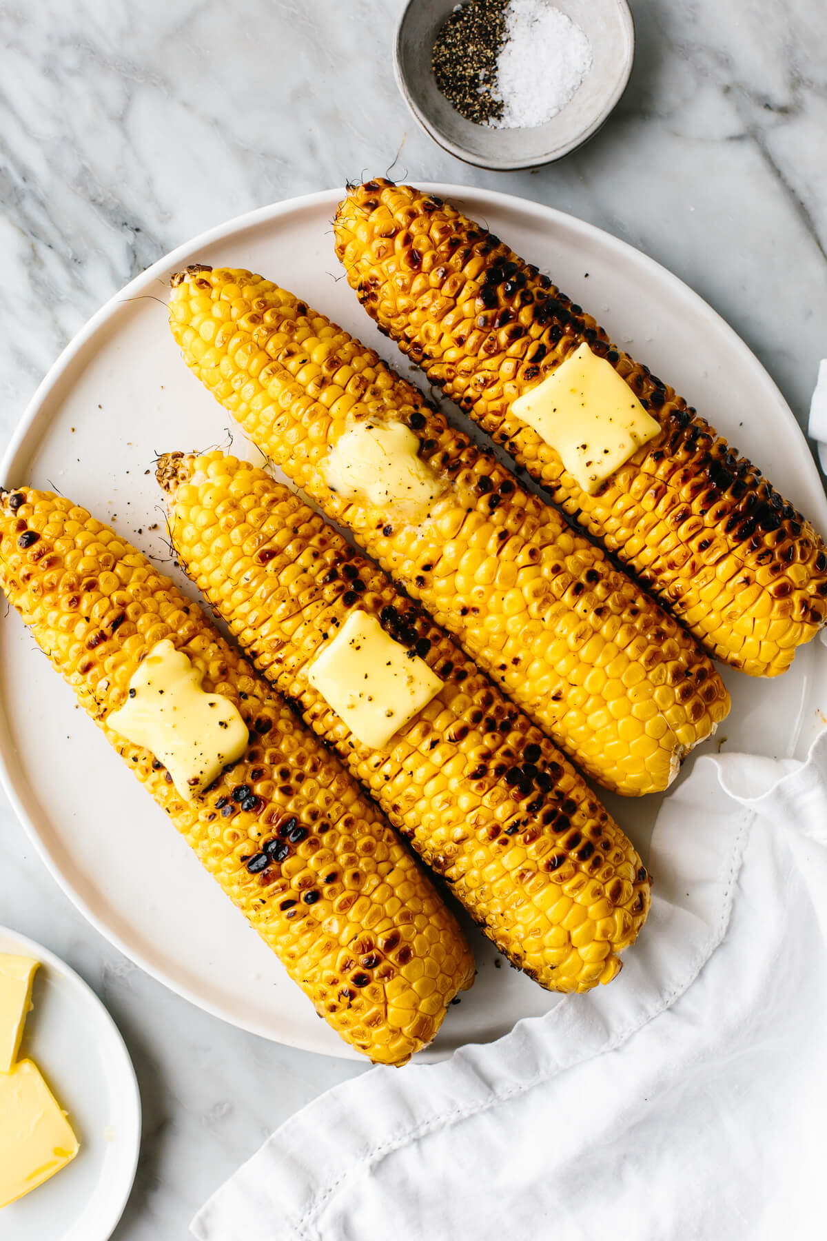 Grilled corns on the cobb on a white plate next to butter.