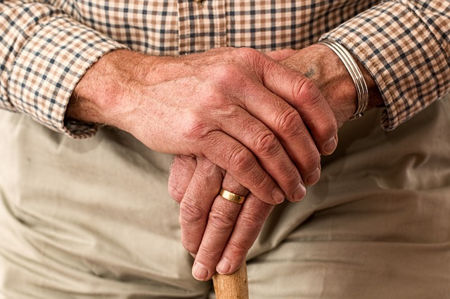 Close up of an elderly man's hands held over teh top of a walking stick. He has a gold wedding ring and is wearing light beige rousers and a brown checked shirt.