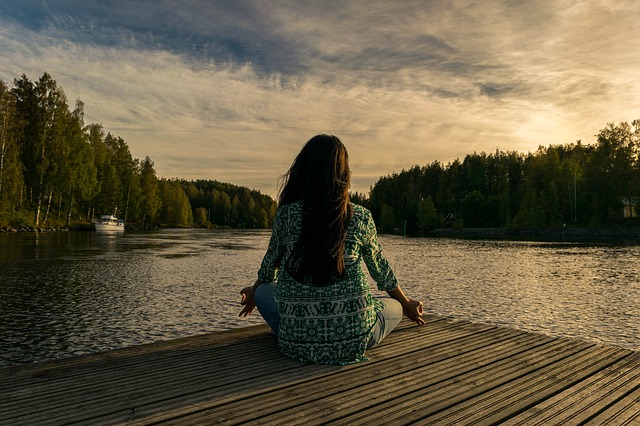 Long haired brunette lady sat crosslegged at the end of a pier overlooking a lake practising mindfulness