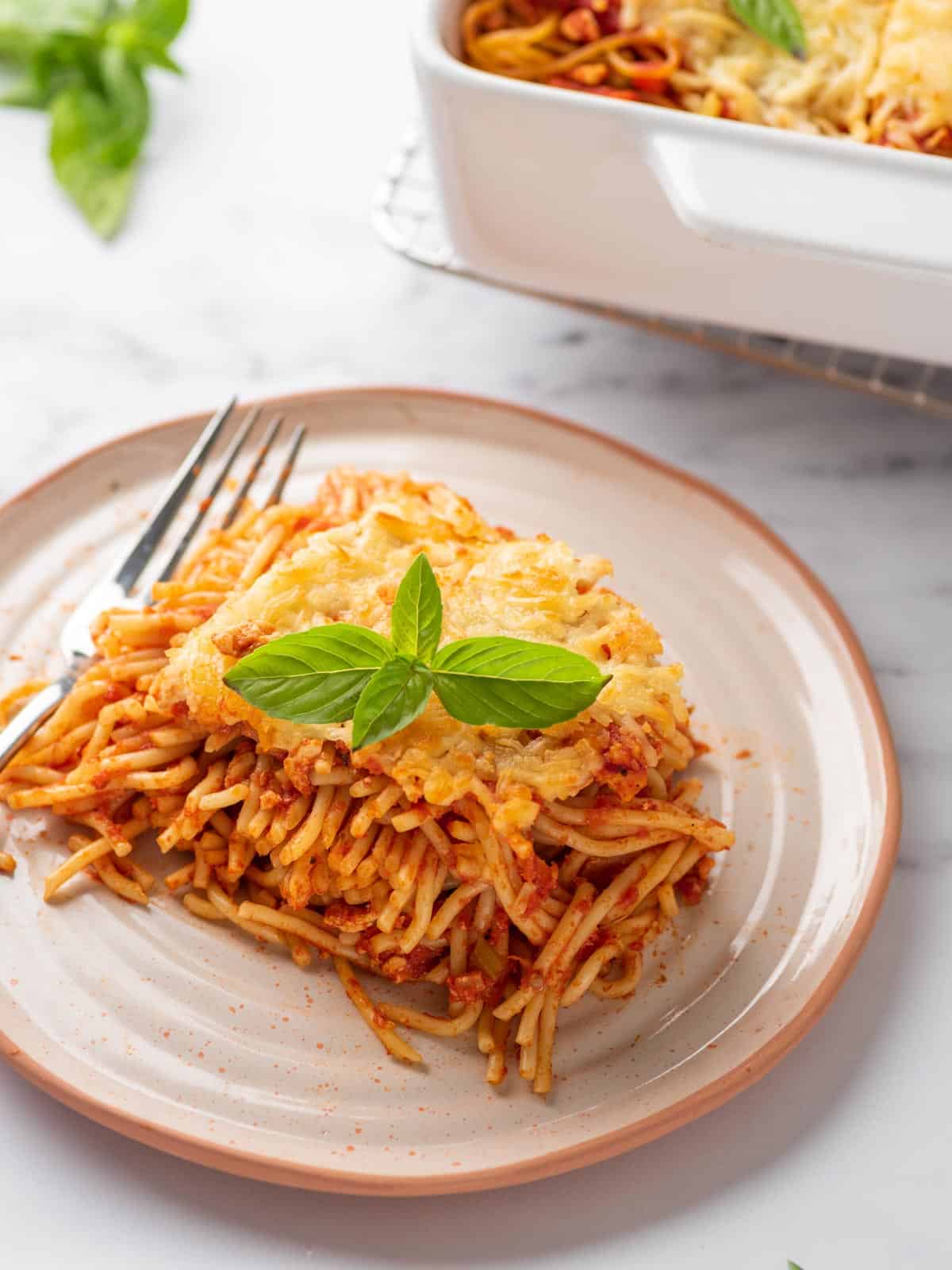 portion of baked spaghetti pasta served on a plate and topped with basil leaves.