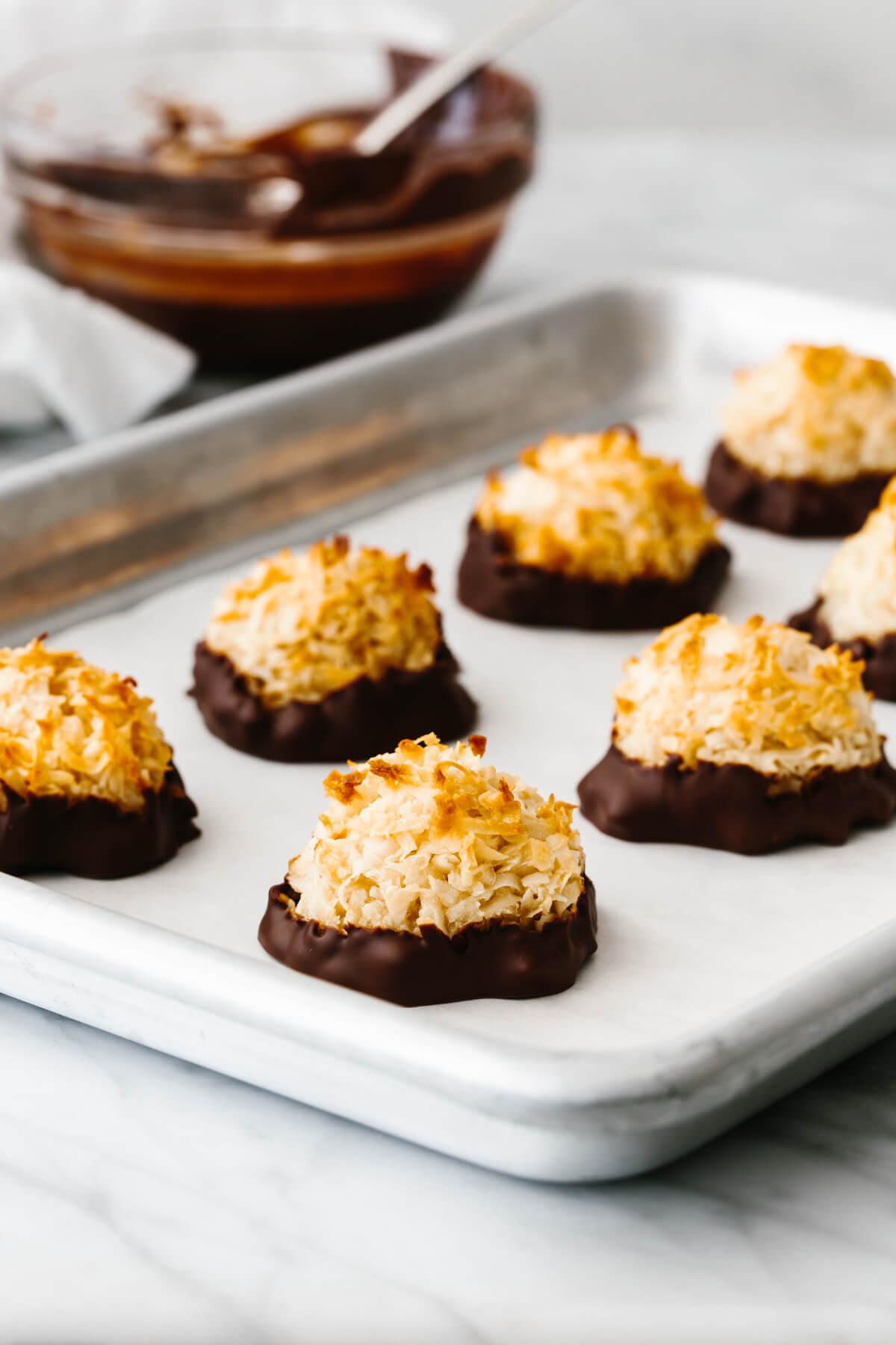 Coconut macaroons on a baking sheet next to a bowl of chocolate.
